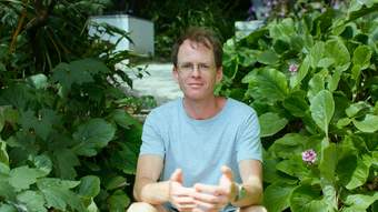 A person wearing a white t-shirt and glasses sitting among many green plants