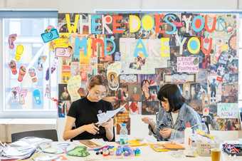 Two people sit working at a table covered in craft materials; behind is a wall collaged with text and images, including large lettering saying: Where does your mind take you?